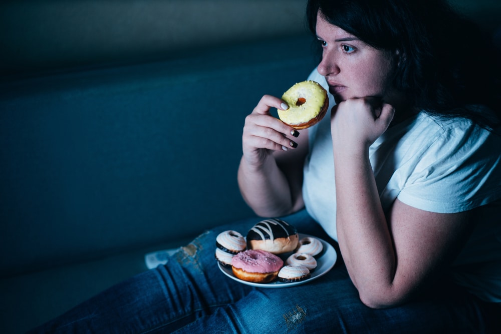 Stressed woman eating a plate of doughnuts in front of the TV due to stress and high cortisol.