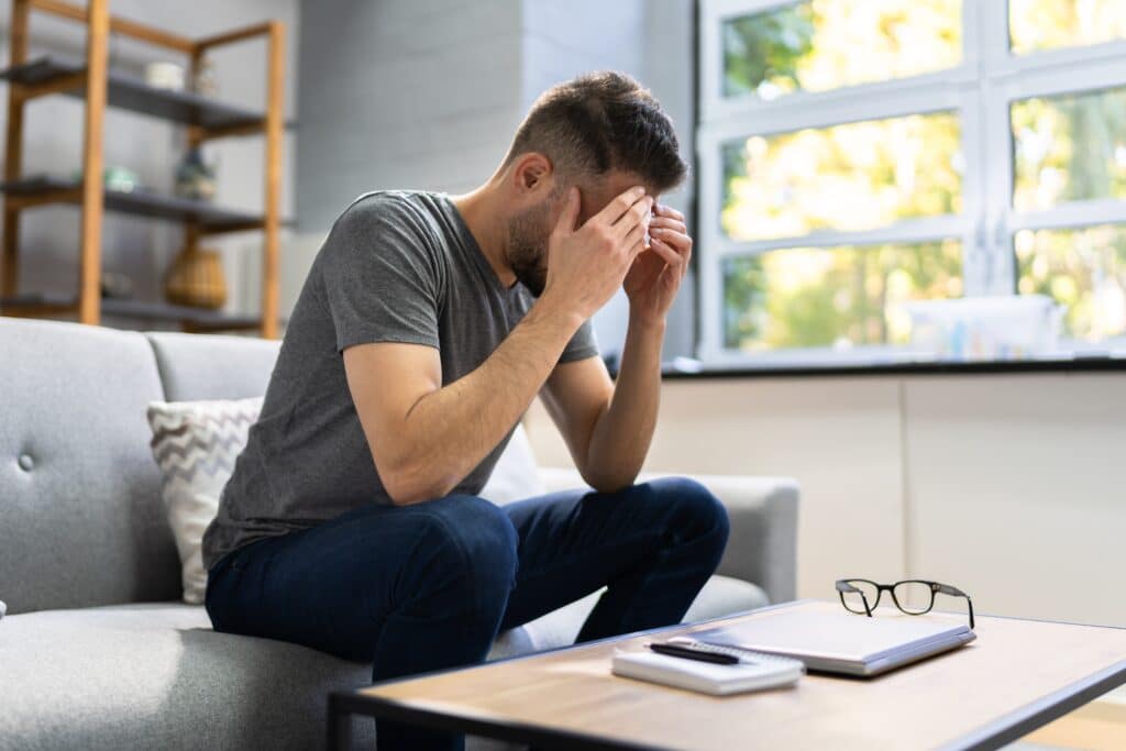 Stressed man sitting on couch and holding is face, feeling the effects of chronically high cortisol