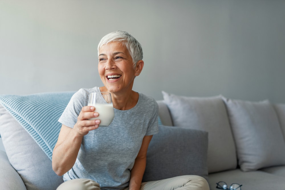 Smiling older woman relaxing on a couch with a glass of milk, representing the importance of maintaining bone health during menopause.