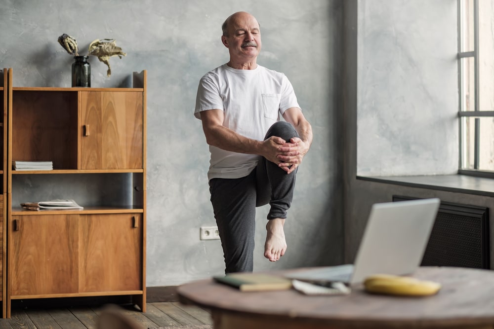 Older man doing a balance exercise indoors with one knee raised, following a workout on a laptop.