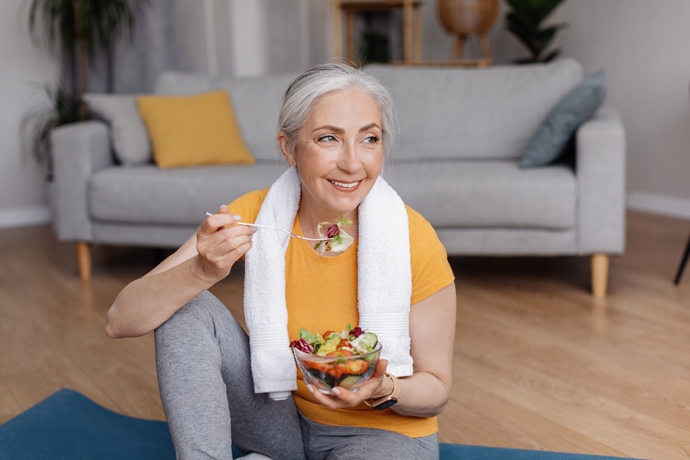 Older woman eating a healthy salad at home after a workout, smiling and relaxed