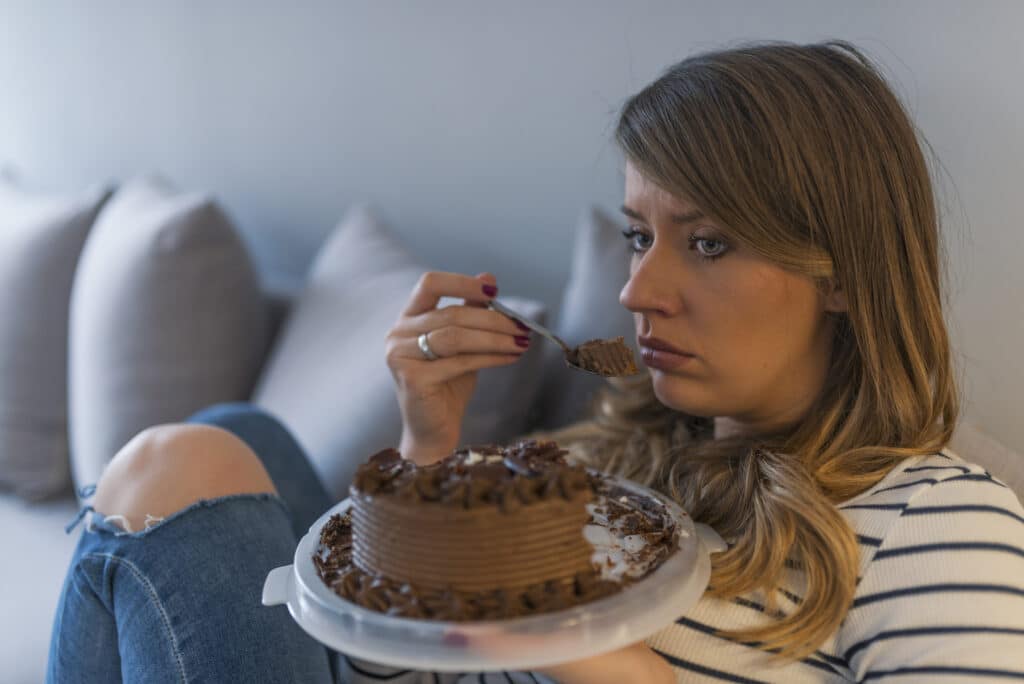 Young woman sitting on couch looking sad and eating a chocolate cake